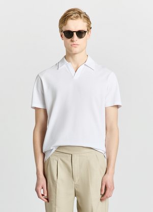 Young man with blonde hair wearing black sunglasses, a white short-sleeve polo shirt, and beige pleated trousers against a plain background.