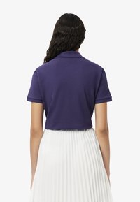 Camisa polo púrpura de manga corta, con un cuello clásico y un diseño corto, combinada con una falda blanca con pliegues. Estilo minimalista.