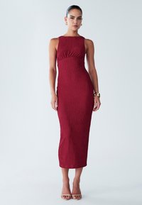 Ženska, oblečena v brezrokavno, teksturirano, oprijeto bordo midi obleko z zlatimi uhanami, zapestnico in bež čevlji z odprtimi prsti, stoji na belem ozadju.