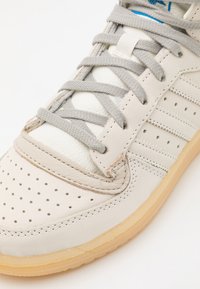 Light grey athletic shoe with perforated leather upper, cotton laces, and rubber sole; features flat grey laces and a padded tongue.