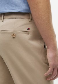 Nahaufnahme einer Person, die beige Hose mit einer geknöpften Gesäßtasche und einem kleinen rot-weiß-blauen Logo in der Nähe der Tasche trägt.