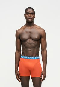 Μυώδης άνδρας που φοράει πορτοκαλί μποξεράκια Björn Borg με μπλε ζώνη, στέκεται μπροστά σε απλό φωτεινό φόντο, κοιτάζοντας προς τα εμπρός.