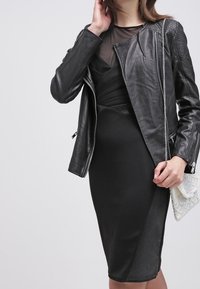 Svart lädermotorcykeljacka med silverdragkedjor, lager på en figursydd svart klänning med genomskinlig halsringning. Håller en texturerad vit väska.