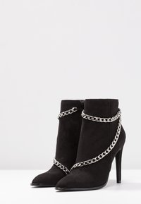 Bottines en daim noir avec bout pointu, talon stiletto et un accent de chaîne argentée entourant la tige, dotées d'une fermeture éclair.