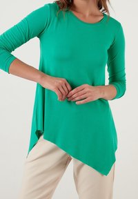 Grön långärmad topp i ett mjukt och stretchigt material, med en asymmetrisk nederkant och rund halsringning, tillsammans med beige byxor.