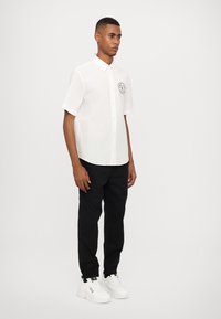 Chemise blanche à manches courtes avec col boutonné, ornée d'un logo circulaire sur la poitrine, associée à un pantalon noir et des baskets blanches.