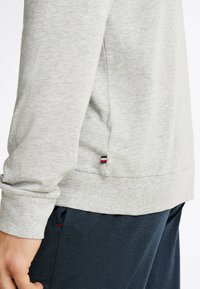 Camisa de manga larga en gris claro hecha de un tejido suave, con una abertura lateral y una pequeña etiqueta de marca con acentos en rojo, blanco y azul cerca del dobladillo.