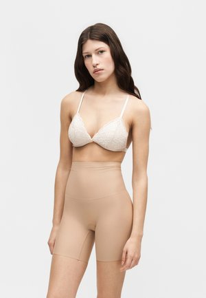 Ung kvinde med langt brunt hår iført hvid blonde-bh og nudefarvede højtaljede formgivende shorts mod en ensfarvet baggrund.