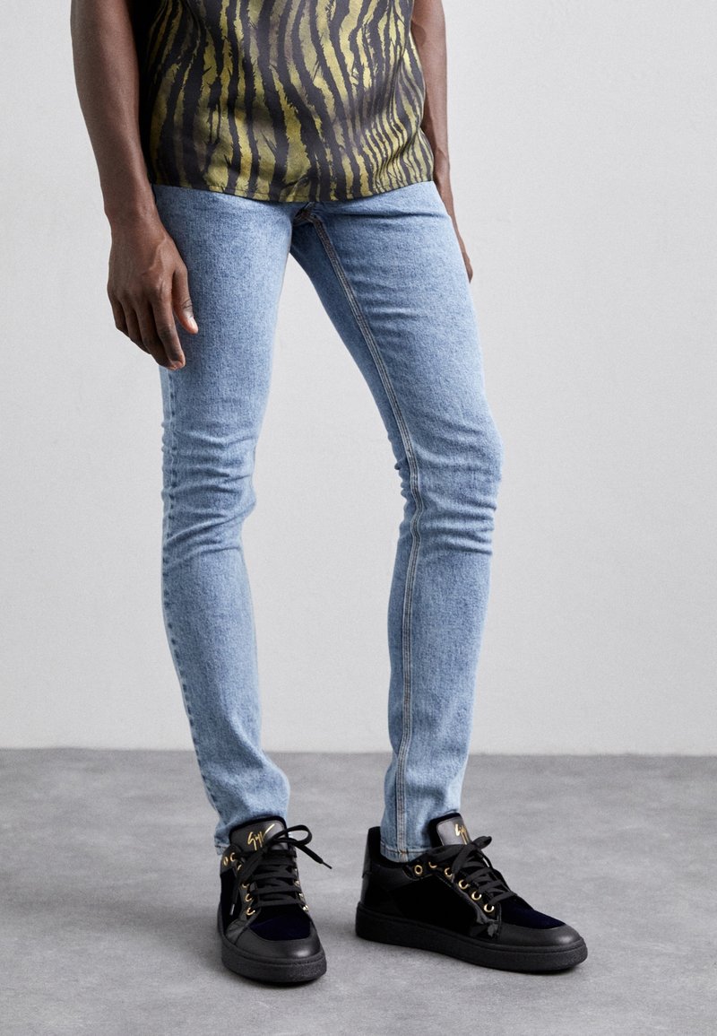 Jeans de corte ajustado en un azul claro combinados con zapatillas negras con detalles dorados y un material texturizado, llevadas con una blusa oscura de estampado.