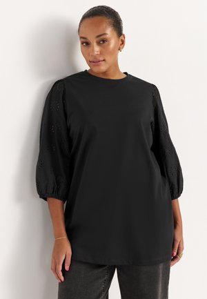 Femme aux cheveux foncés attachés en arrière, portant un chemisier noir avec des manches bouffantes brodées et un pantalon sombre, debout contre un mur blanc.