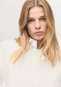 Maglione bianco a treccia con scollo rotondo, indossato sopra una camicia bianca con colletto. I capelli biondo chiaro coprono parzialmente la spalla.