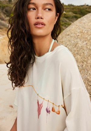 Tânără cu păr lung și creț, purtând o cămașă albă cu un imprimeu reprezentând o sfoară de rufe pe care sunt atârnate costume de baie, în aer liber, aproape de pietre și vegetație.