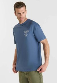 Hombre vistiendo una camiseta gráfica azul con el texto "The Fries" y pantalones verdes, mirando hacia abajo y ligeramente de lado contra un fondo blanco.