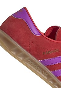 Czerwony zamszowy sneaker z fioletowymi skórzanymi akcentami, trzema równoległymi paskami, teksturowaną podeszwą, złotym napisem "HAMBURG" oraz logo adidas.