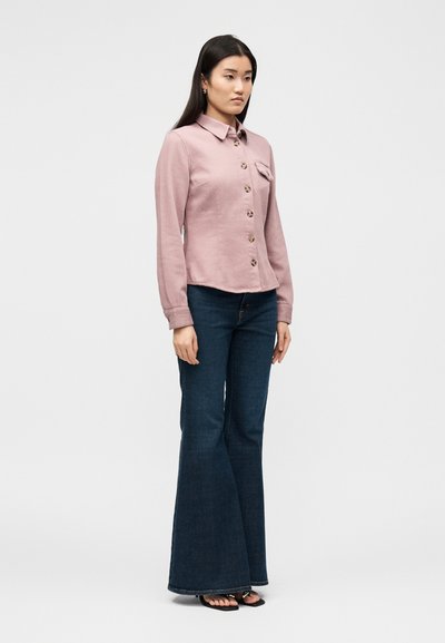 Γυναίκα όρθια, φοράει μια πουκάμισο σε σκόνη ροζ απόχρωση με τσέπη στο στήθος, σκούρα μπλε καμπάνα τζιν και μαύρα σανδάλια με ανοιχτά δάχτυλα.