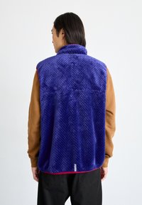 Purple fleece vest with a textured pattern, featuring a high collar and red accents, worn over a brown long-sleeve shirt.