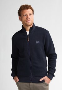 Granatowa kurtka zapinana na zamek z teksturowanej tkaniny, z kołnierzem typu stójka i zamkiem z przodu. Posiada boczne kieszenie oraz metkę na piersi.