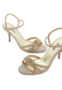 Sandales à talons hauts dorées avec une tige en tissu texturé, des sangles ornées, un design noué et une doublure intérieure métallique.