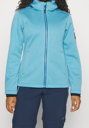 Vrouw met een lichtblauwe capuchontrui met rits en donkerblauwe broek, staand met ontspannen armen langs het lichaam tegen een effen achtergrond.