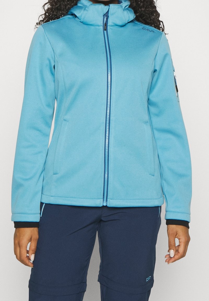 Femme portant une veste à capuche zippée bleu clair et un pantalon bleu foncé, debout avec les bras détendus le long du corps devant un fond uni.