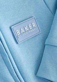 Lichtblauwe stof met een gestikt rechthoekig label waarop "BAKER BY TED BAKER" in het wit staat, met een textuur van gebreide stof en een opgerolde mouwomslag.