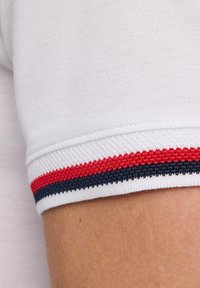 Biała koszula z krótkim rękawem z teksturowanym kołnierzykiem w czerwone i granatowe poziome paski. Zbliżenie na detal rękawa.