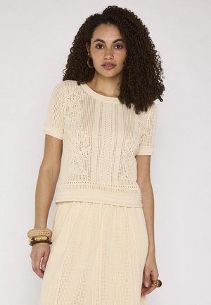 Donna con capelli ricci che indossa un top lavorato a maglia crema a maniche corte e una gonna abbinata, accessoriata con braccialetti e orecchini a cerchio.