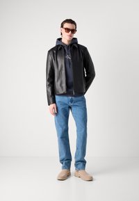 Black leather jacket with a zip front, navy hoodie underneath, blue jeans, and beige slip-on shoes; model wearing brown sunglasses.