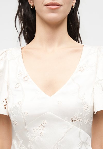 Γυναίκα που φοράει λευκό φόρεμα σε σχήμα V με κεντήματα λουλουδιών και κοντά φουσκωτά μανίκια, χρυσούς κρίκους στα αυτιά και φυσικό μακιγιάζ.