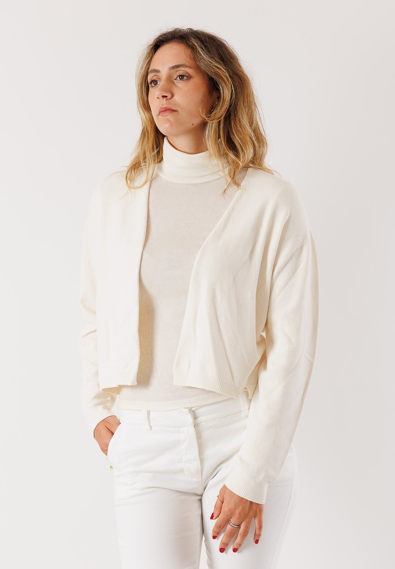 Cardigan accorciato color crema con maniche lunghe, materiale leggero, design aperto davanti, abbinato a un dolcevita aderente e pantaloni bianchi.