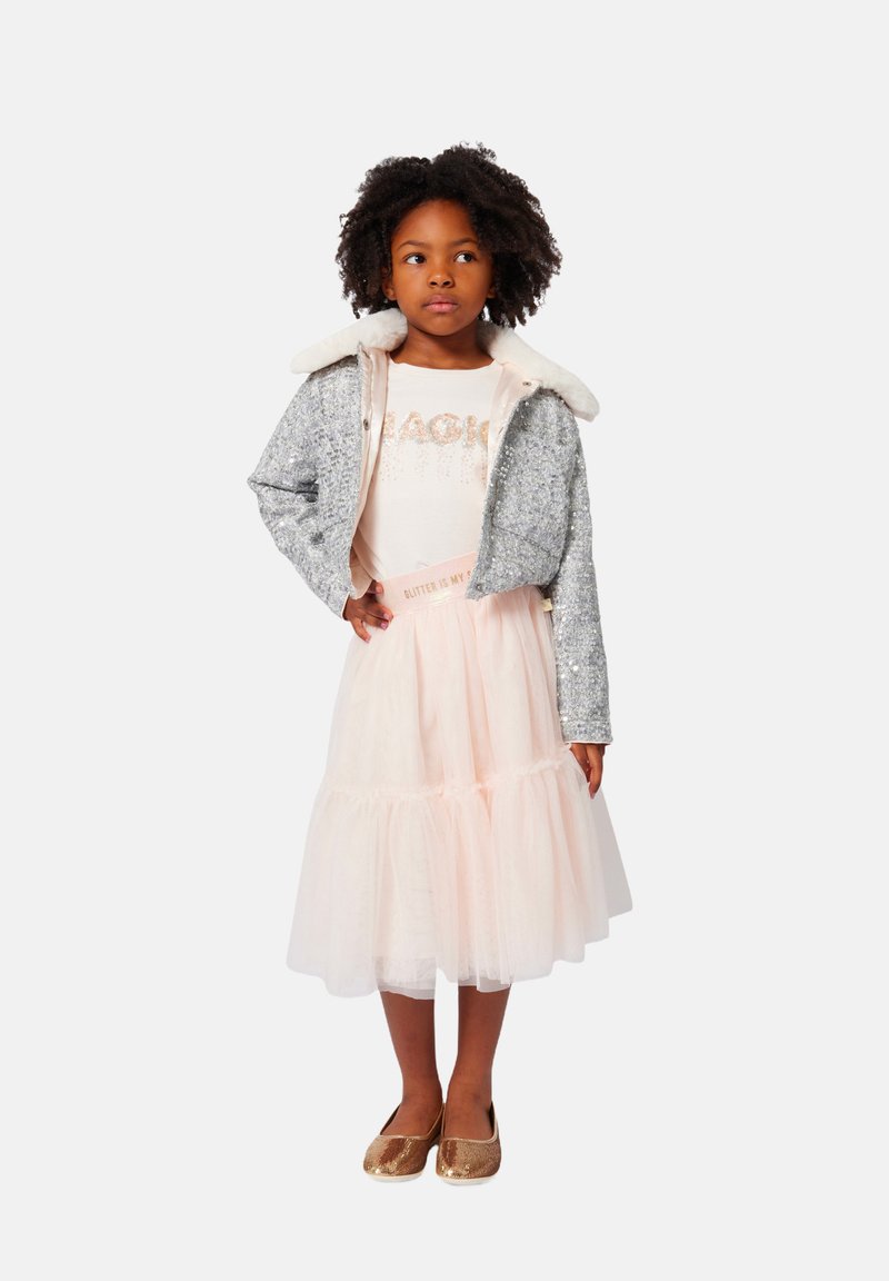 Veste en sequins argentés avec une doublure intérieure blanche, jupe en tulle rose clair avec volants, et chaussures dorées brillantes. Le haut présente un texte pailleté.