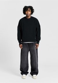 Black knitted sweater with oversized fit, ribbed texture, and wide sleeves, paired with loose-fitting dark gray jeans and light sneakers.