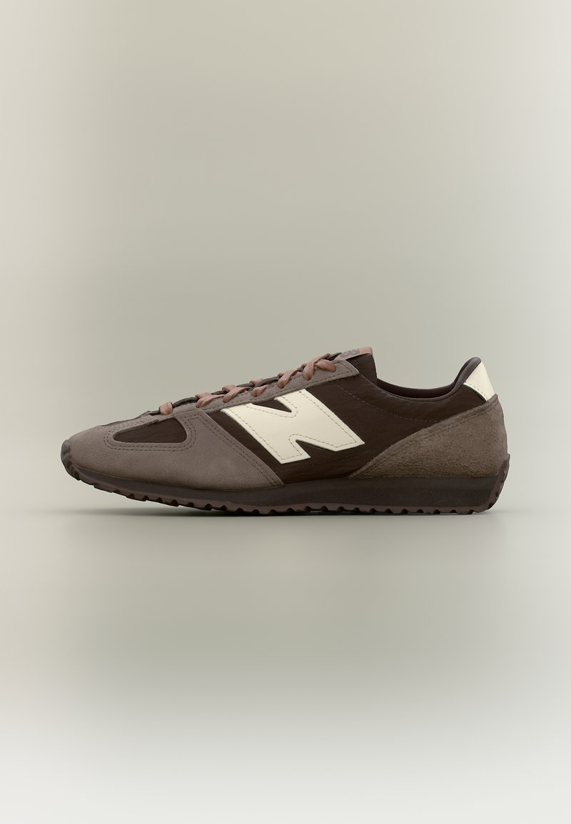 Καφέ και γκρι αθλητικό παπούτσι χαμηλού τύπου με μπεζ λογότυπο "N" στο πλάι, κορδόνια για κλείσιμο και υφή στην σόλα, εμφανιζόμενο σε προφίλ σε ουδέτερο φόντο.