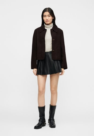 Γυναίκα που φοράει καφέ σακάκι με κουμπιά, ριγέ ζιβάγκο, μαύρη πλισέ φούστα και μαύρες μπότες μέχρι τη μέση της γάμπας, στέκεται μπροστά σε ένα μονόχρωμο φόντο.