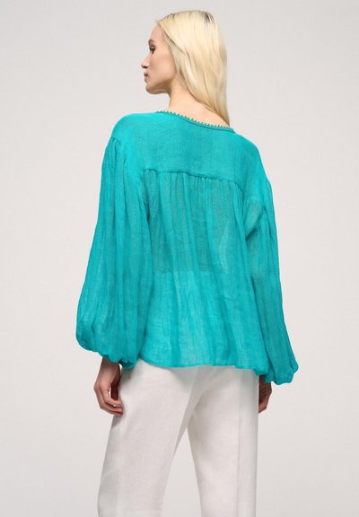 Bluză din in turquoise, cu mâneci lungi, strânse și pliuri la spate; are un decolteu rotund cu un bordaj texturat. Purtați cu pantaloni albi.