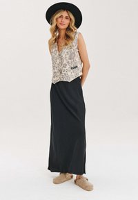 Béžový bezrukávový top s paisley vzorom, čierna maxi sukňa a béžové slip-on topánky. Modelka má na sebe čierny klobúk s širokým okrajom.