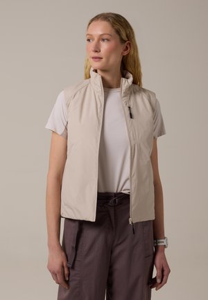 Mujer rubia con chaleco beige sin mangas con cremallera sobre camiseta blanca y pantalones morados, mirando ligeramente hacia la izquierda.