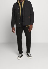 Svart canvasjacka med knäppningar och bröstfickor, lager på lager över en gul knäppskjorta, kombinerad med svarta jeans och svarta sneakers.