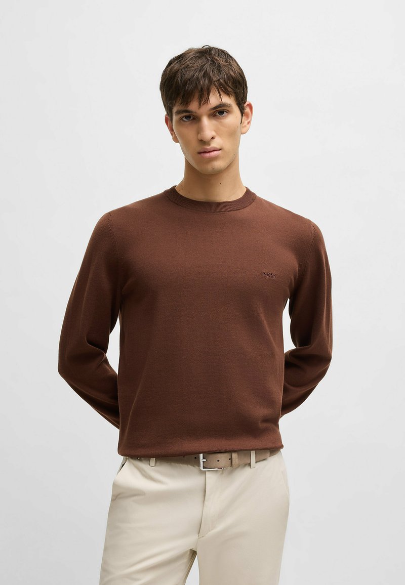 Brown knit sweater made of textured fabric, featuring a round neckline and long sleeves. Small logo detail on the chest, paired with light pants.