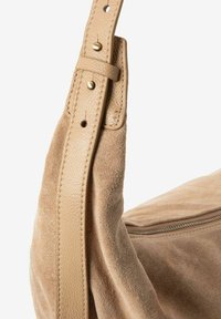 Sac à main en suede beige avec une sangle en cuir lisse ornée de rivets métalliques et une fermeture éclair, mettant en avant une texture douce et des tons neutres.