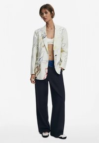 Weißes Blazer mit bunten Blumenmustern, kombiniert mit einem weißen Crop-Top und locker sitzenden dunkelblauen Hosen. Mit weitem Schnitt und kontrastierenden schwarzen Schuhen.