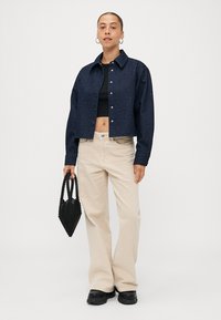Navy denim cropped jacket met een knoopsluiting over een zwarte top, gecombineerd met beige ruime broek en een zwarte beaded handtas. Zwarte schoenen.