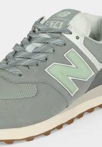 Γκρι και λευκό αθλητικό παπούτσι New Balance 574 με σχέδιο στα γκρι κορδόνια, ανοιχτά πράσινο λογότυπο "N" και υφασμάτινο και σουέντ επάνω μέρος με υφή.