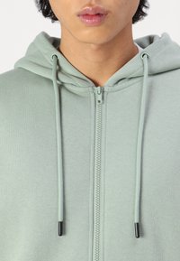 Svijetlozelena zip-up dukserica napravljena od mekanog materijala, s prednjim patentnim zatvaračem, kapuljačom s vezicom i teksturiranim materijalom s glatkom završnom obradom.