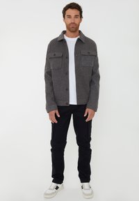 Grå flanneljakke med krave og to brystlommer, iført over en hvid T-shirt, parret med sorte bukser og hvide sneakers.