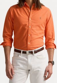 Orange skjorta med knappar och långa ärmar samt en enda logga på bröstet, kombinerad med vita denimbyxor och ett brunt bälte.