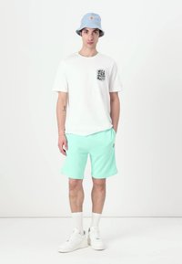 T-shirt branco de manga curta com um bolso de logótipo preto, chapéu de balde azul-claro, calções verdes-mint, ténis brancos e meias brancas de cano curto.