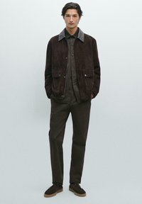 Bruine suède jas met een grijze kraag, daaronder een knoopshirt en donkere broek, gecombineerd met bruine schoenen. Eenvoudig en gestructureerd ontwerp.