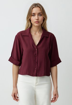 Bordeaux kortekleding met een kraagontwerp, voorzien van knopen aan de voorkant en een korte snit, gecombineerd met witte broeken. Soepele stof.