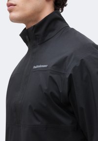 Κοντινό πλάνο ενός ατόμου που φοράει μαύρο μπουφάν με ψηλό γιακά και φερμουάρ, εμφανίζοντας το λογότυπο «Peak Performance» στο στήθος.